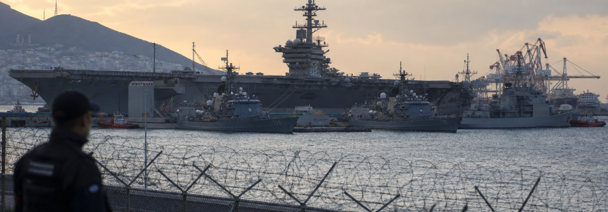 A naval harbor at dusk with a large aircraft carrier berthed behind security fencing.