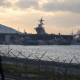 A naval harbor at dusk with a large aircraft carrier berthed behind security fencing.