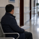 A man sitting alone on a hospital corridor bench holding a folder of documents.