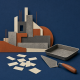 Cut-paper building shapes beside a plastering trowel, an empty tray and blank paper slips on an indigo background.
